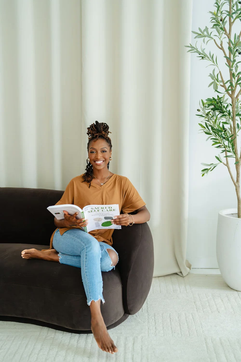 Brittany Ellis seated in a cozy chair, smiling and holding her book Sacred Self Care